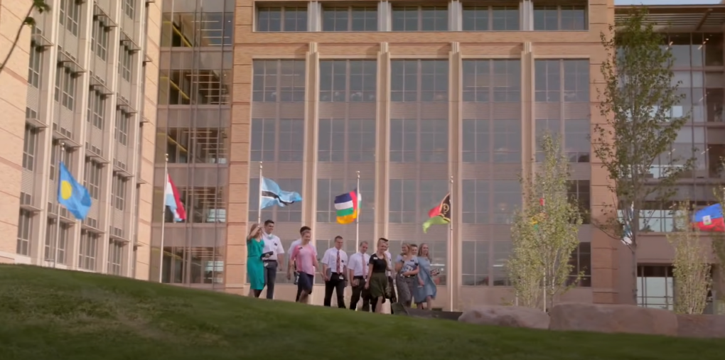 People walking outdoors with a building and various flags in the background